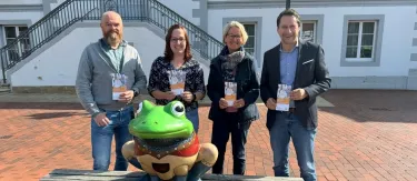 Vier Personen stehen hinter einer großen Froschfigur.